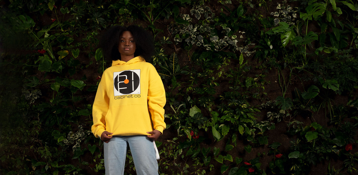 Person wearing a yellow hoodie with the Hifi Cabinet Co logo on the front. In the background is a wall of greenery