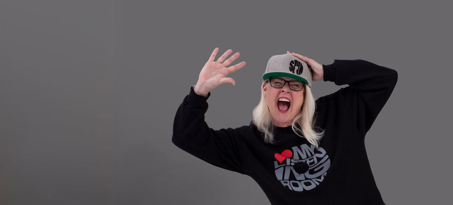 Woman on a grey background wearing a black crewneck with the words 'Love my listening room' in grey. The word love is shown as a red heart instead of the actual word